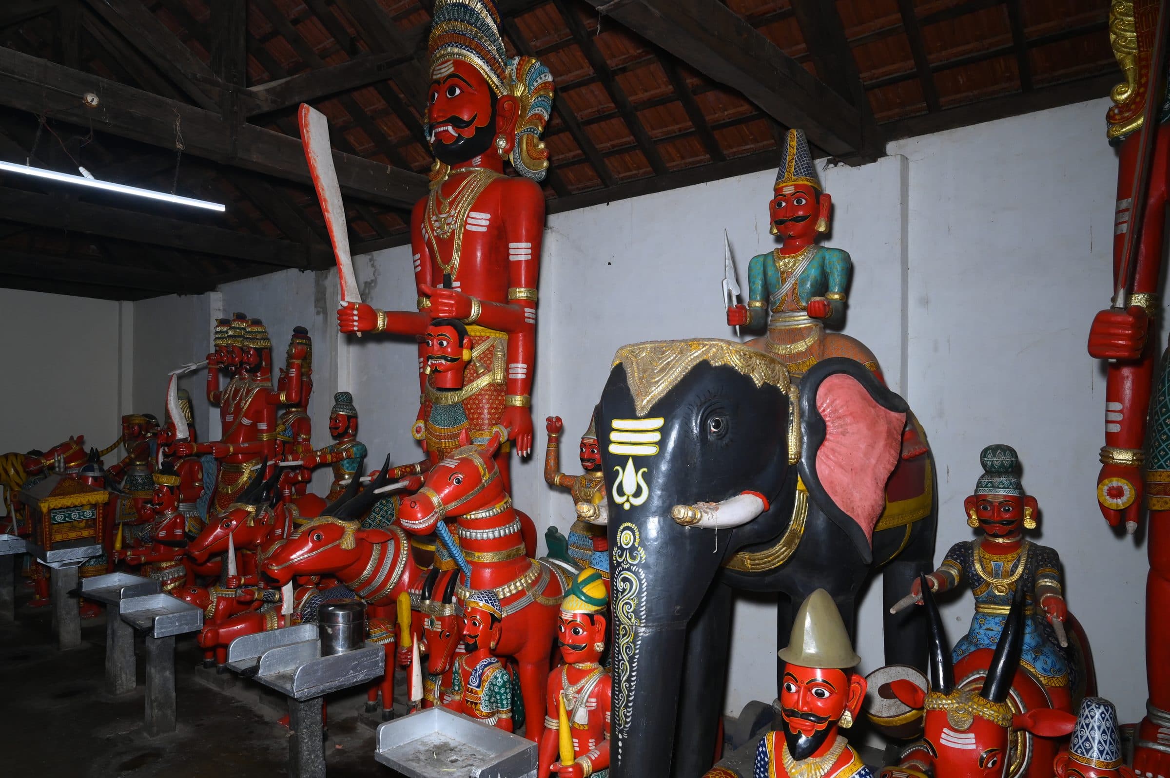 Inside the hall where the wooden figures stand: a tall red figure holding a sword beside a large black elephant mount, further painted figures ranged in rows behind them on low platforms, under a tiled roof carried on timber beams.