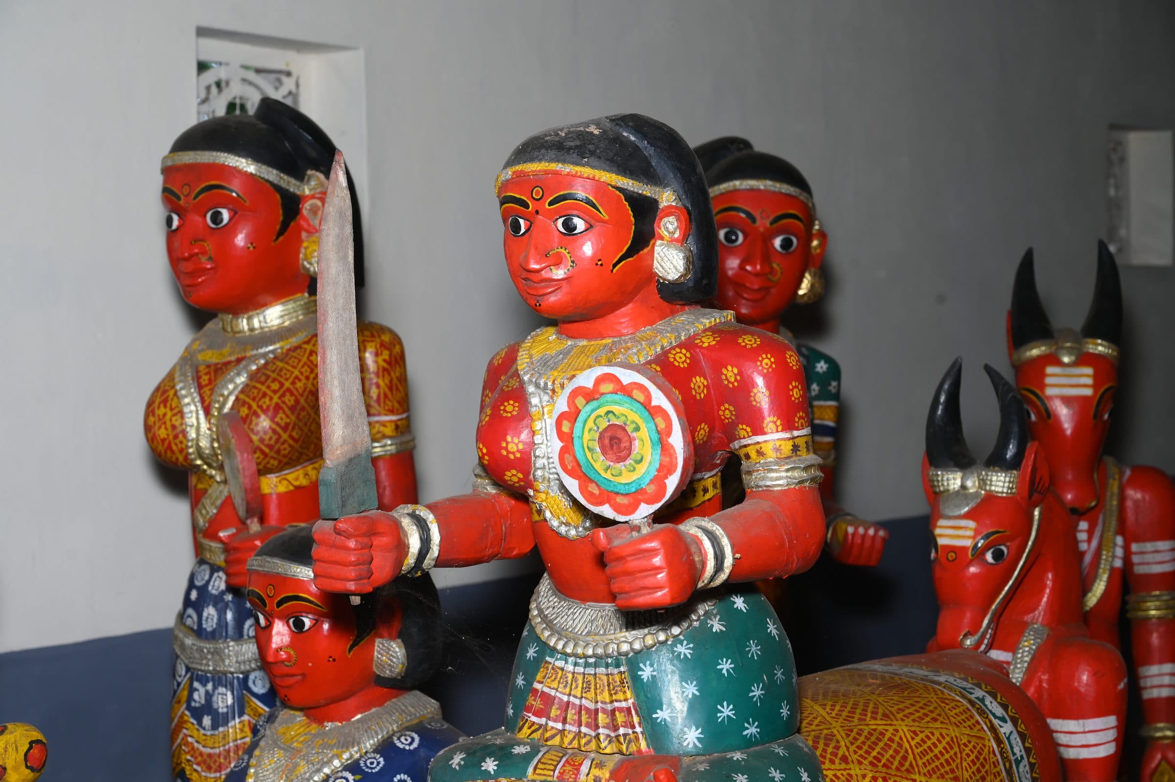 Close view of carved wooden figures painted red, with black hair, patterned garments and silver ornaments — one holding a sword, another a decorated disc — with a red wooden bull beside them.