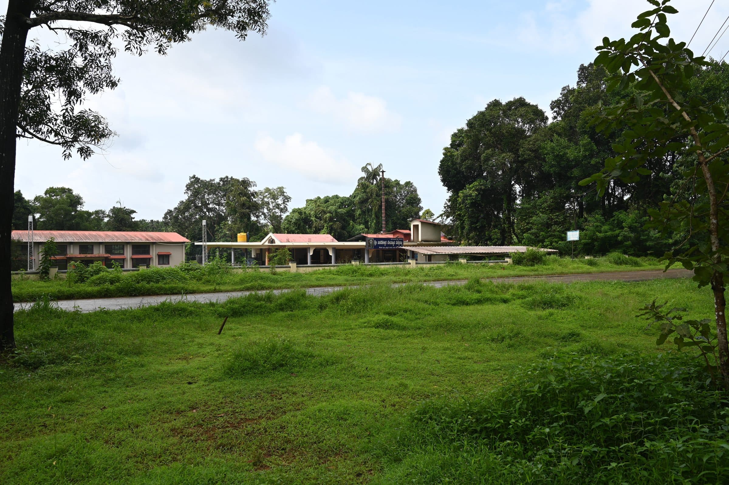 Open green ground in front of the temple: grass and low bushes in the foreground, and the long tiled buildings standing among trees along the far edge under a clouded sky.