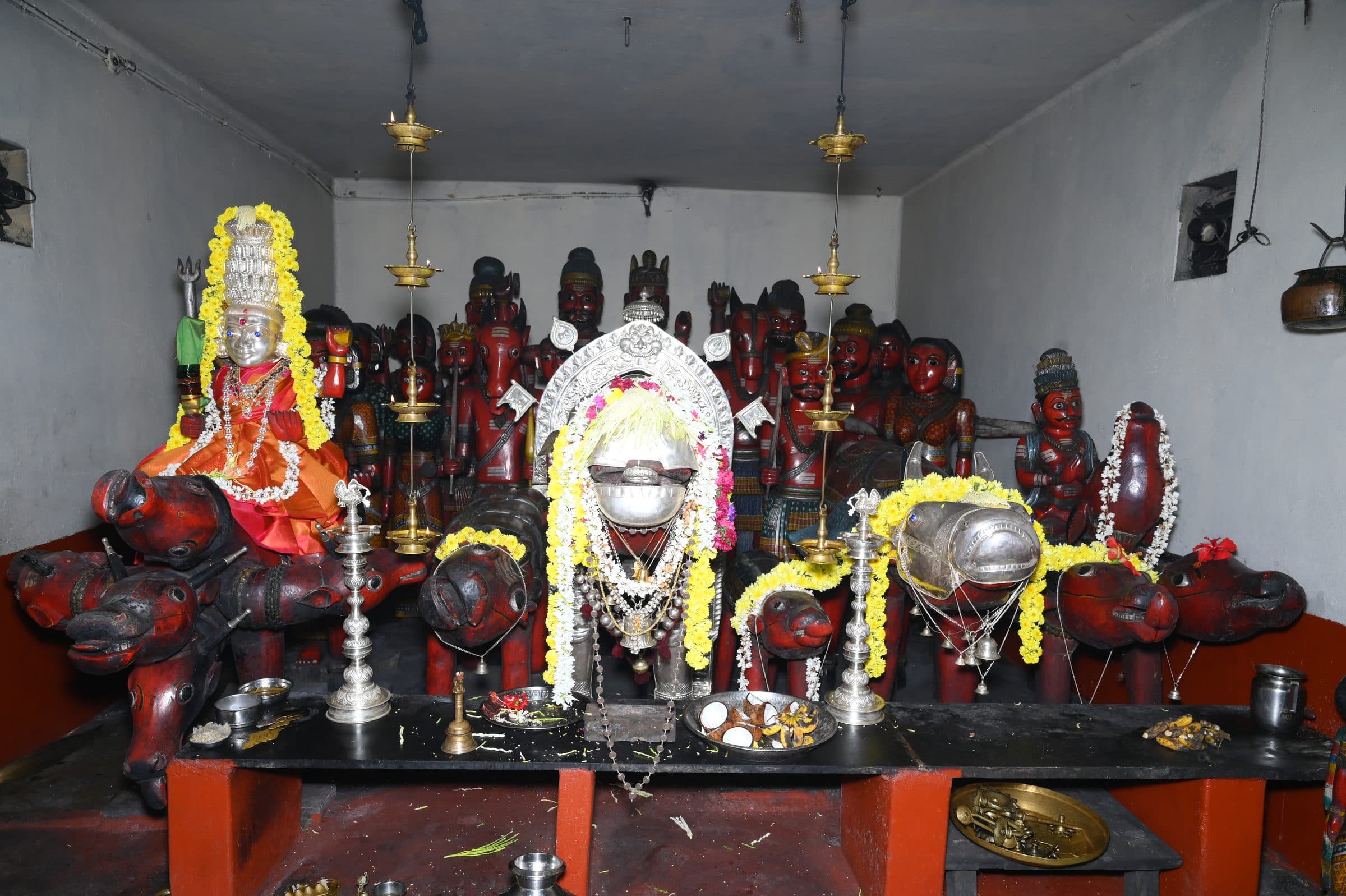 The shrine chamber seen from the front: garlanded silver-covered figures at the centre, rows of red painted wooden figures behind, carved animal mounts along the platform, hanging brass oil lamps and offering vessels in front.