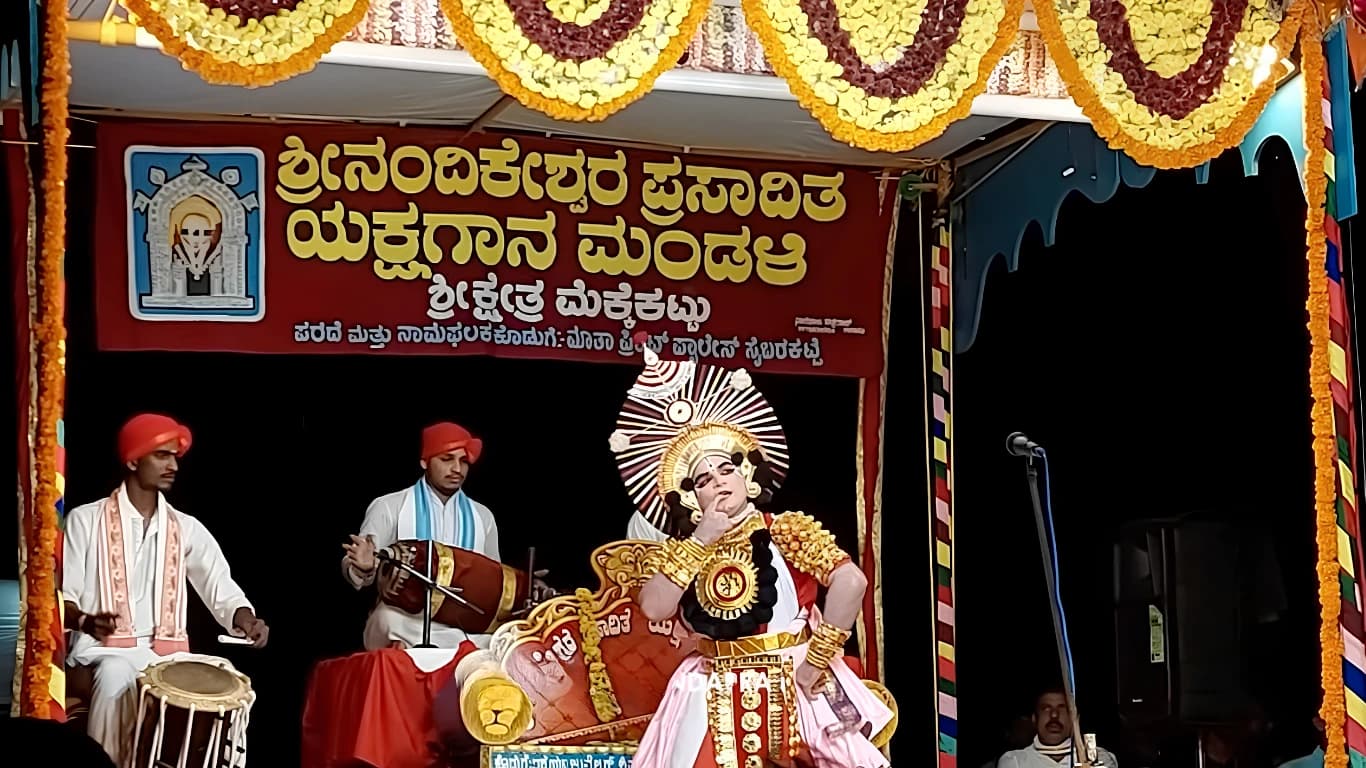 ರಂಗಸ್ಥಳದಲ್ಲಿ ಬಣ್ಣದ ಮುಖವರ್ಣಿಕೆ, ಚಿನ್ನದ ಆಭರಣ ಮತ್ತು ದೊಡ್ಡ ಅಲಂಕಾರಿಕ ಕಿರೀಟ ಧರಿಸಿದ ಯಕ್ಷಗಾನ ಕಲಾವಿದ; ಪಕ್ಕದಲ್ಲಿ ಬಿಳಿ ಉಡುಗೆ ಮತ್ತು ಕೆಂಪು ಪೇಟ ಧರಿಸಿದ ಇಬ್ಬರು ವಾದ್ಯಗಾರರು ಕುಳಿತಿದ್ದಾರೆ; ಮೇಲೆ ಮಂಡಳಿಯ ಕೆಂಪು ಫಲಕ ಮತ್ತು ಚೆಂಡುಹೂವಿನ ಮಾಲೆಗಳ ಸಾಲು.